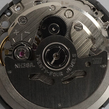 A close-up of a watch movement mechanism with visible gears, bridges, screws, and a clear plastic adjustment knob on a light gray background.