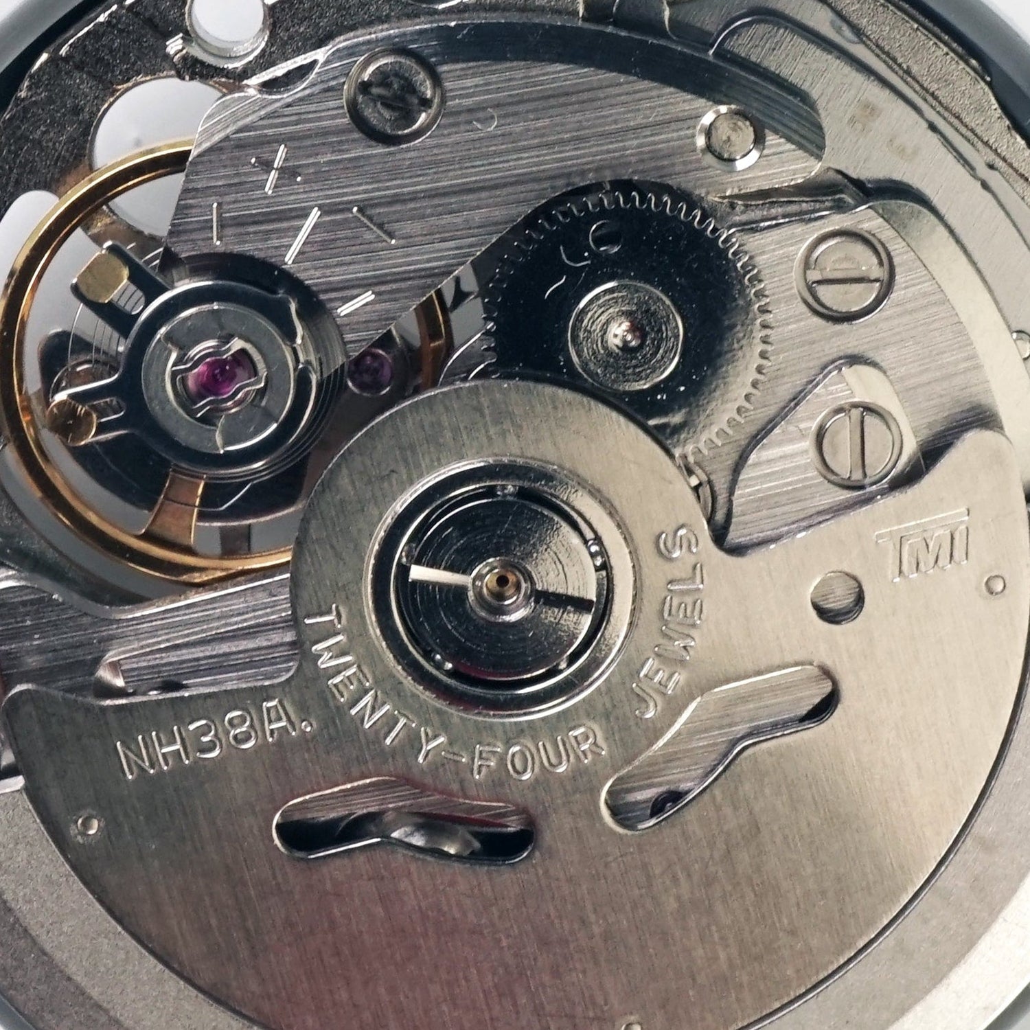 A close-up top-down view of a watch movement or mechanical watch component with visible gears, bridges, and screws, plus a small blue screwdriver handle on the right side, photographed on a plain white background.